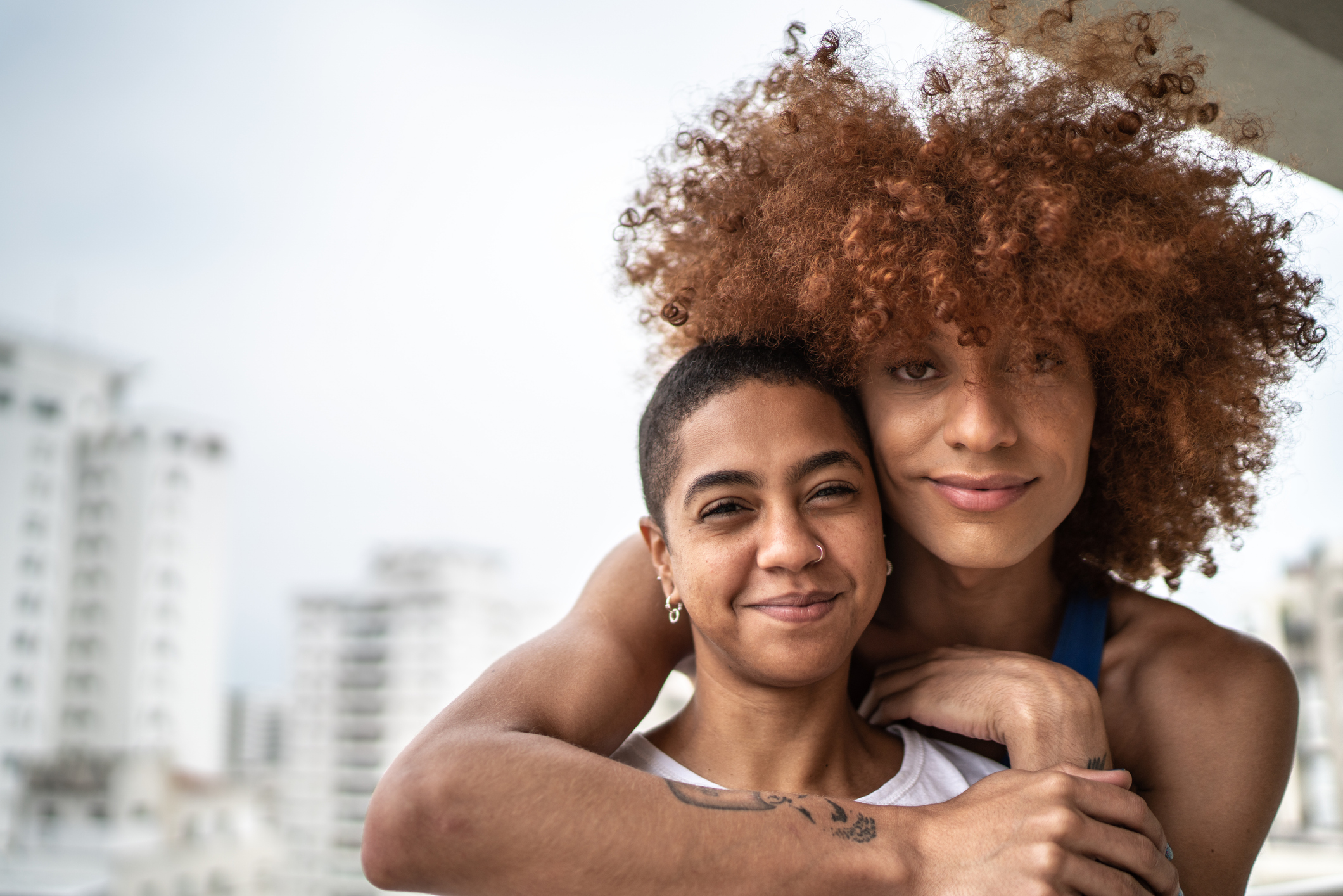 Two brown-skinned trans people hug and look sweetly at the camera. 