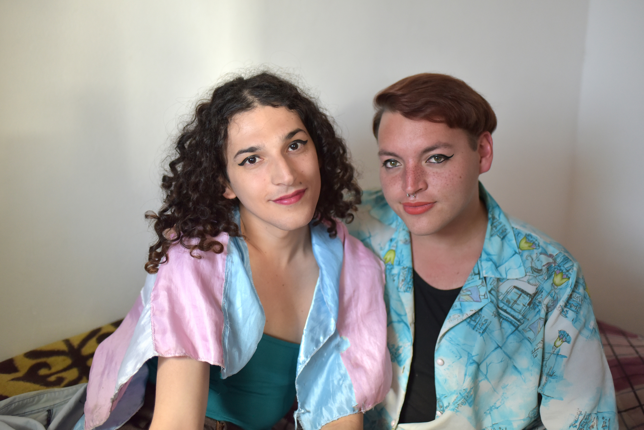 A transfemme person and a nonbinary person wear the trans flag and look confidently at the camera. 