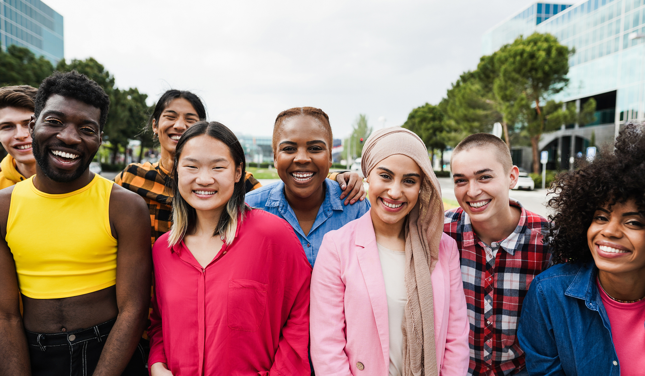 A multi-racial group of people stand together outside and smile in front of the camera
