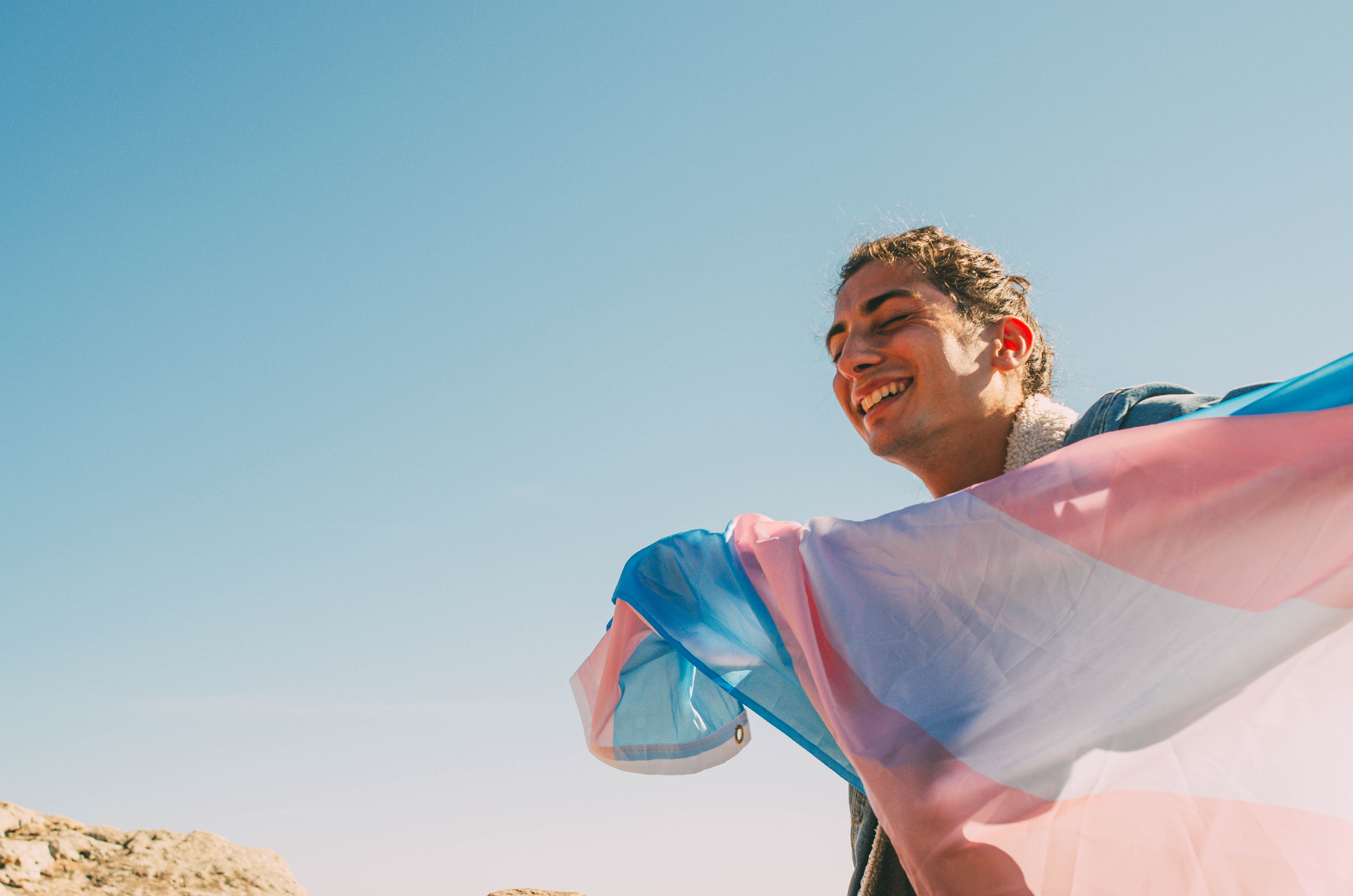 A person with light brown skin wears their hair pulled back. They smile as they hold up the trans flag against the wind. 
