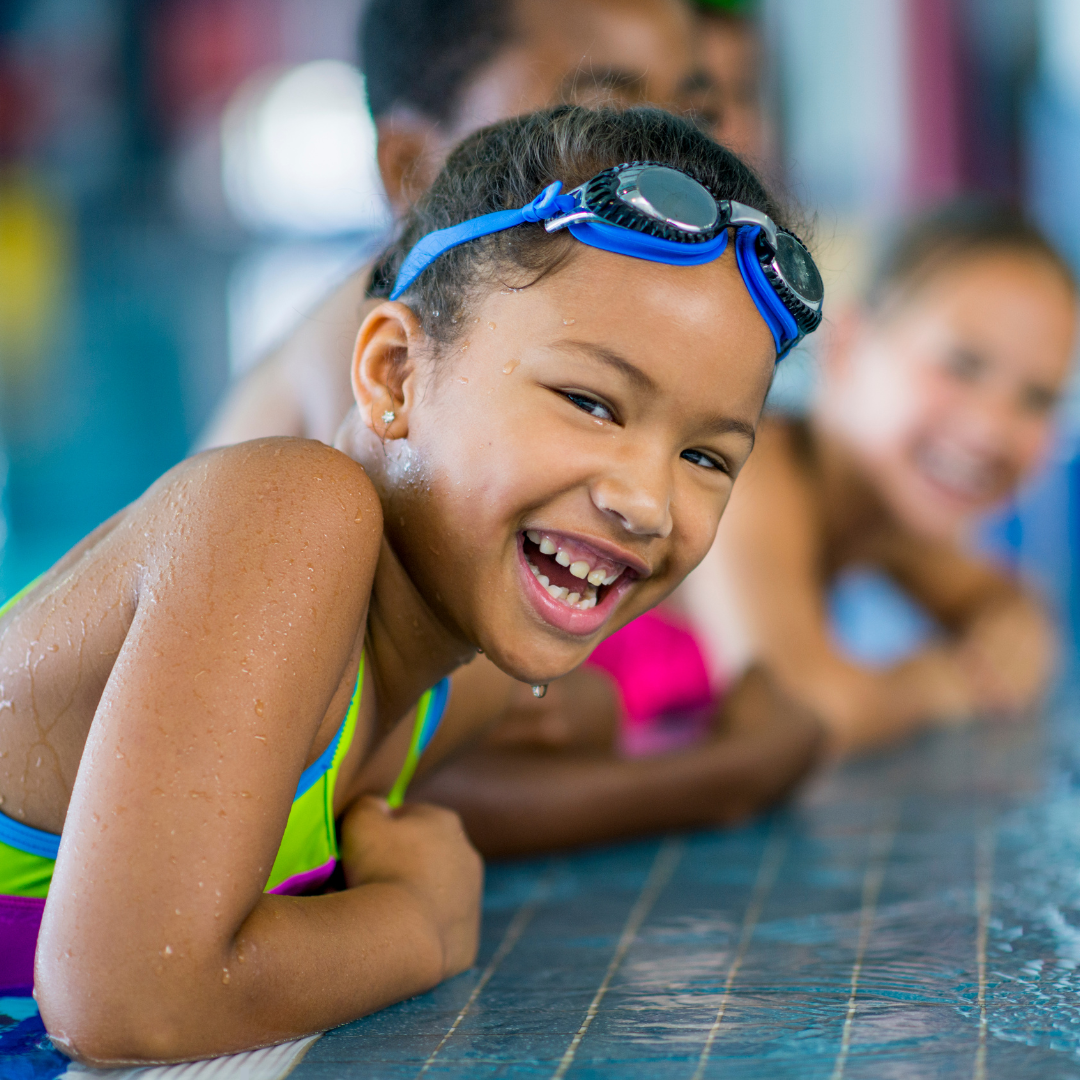 A young Black swimmer smiles at the camera from the edge of the pool. 