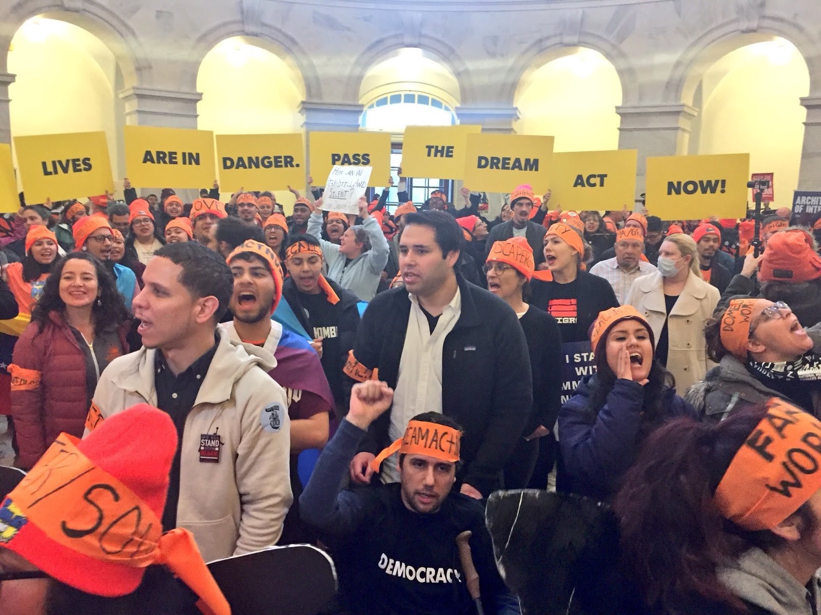 A crowd of protesters, many wearing orange hats and headbands, in the rotunda of the U.S. Capitol. Protesters in the back hold up signs saying "Our lives are in danger. Pass the Dream Act now!"