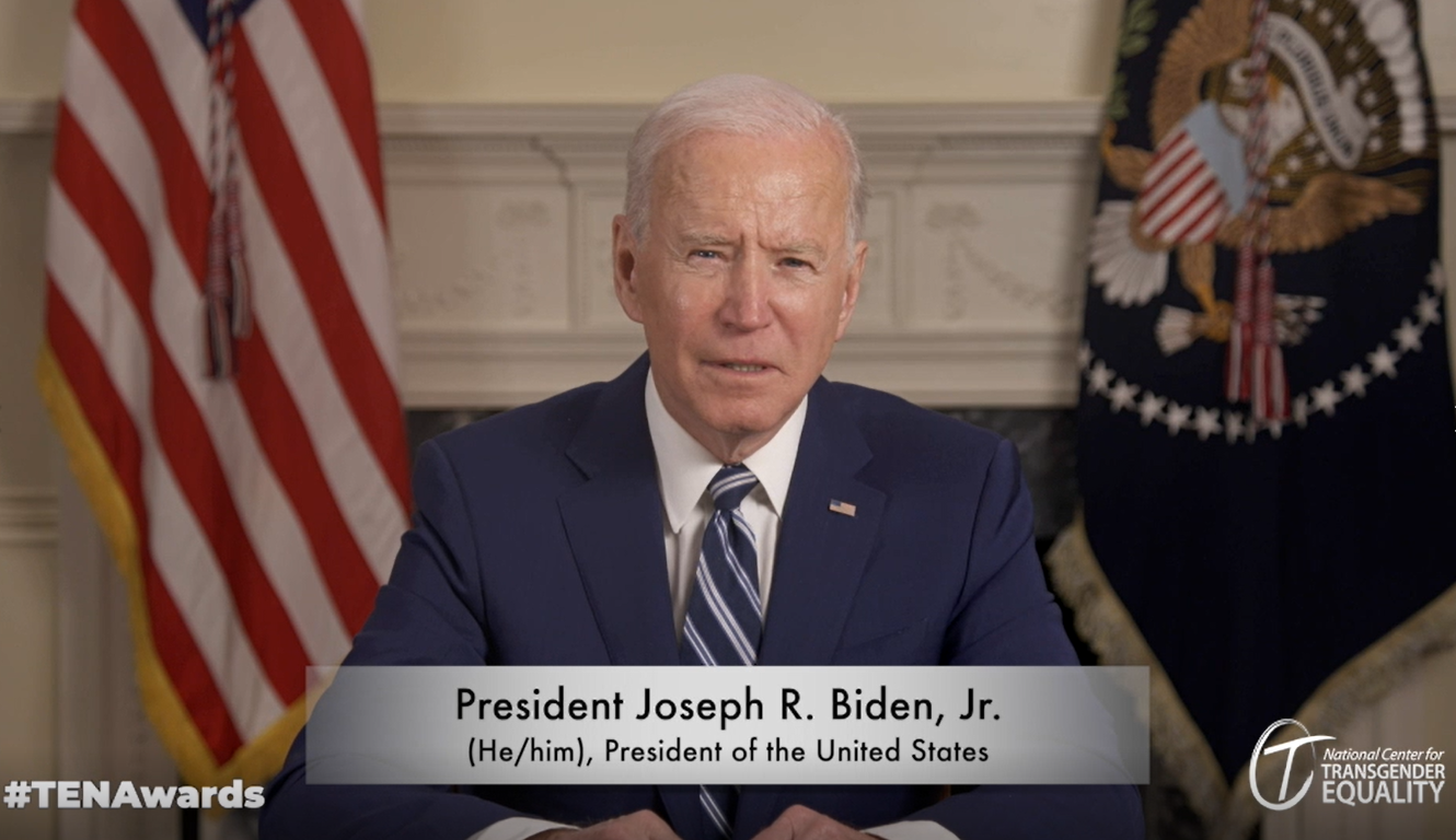 President Biden sitting at desk with digital banner showing he/him pronouns